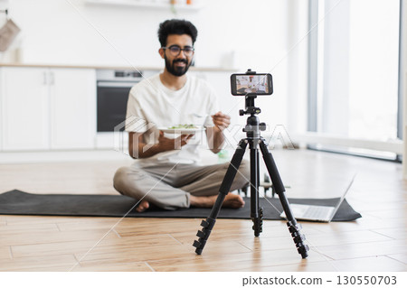 Young adult man sitting cross-legged on yoga mat in modern kitchen recording video blog with smartphone and tripod holding healthy salad plate. Scene suggests health, nutrition, video blogging. Young adult man sitting cross-legged on yoga mat in modern kitchen recording video blog with smartphone and tripod holding healthy salad plate. Scene suggests health, nutrition, video blogging. 130550703
