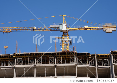 A building under construction has a large crane above it. A construction worker stands on the top level of the structure on a bright day 130552037