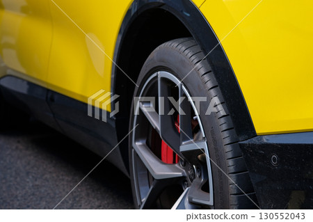 A close-up view captures the wheel, tire, red brake calipers, and section of the bright yellow exterior body of a vehicle parked on the street during the daytime 130552043