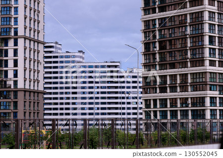 Several modern residential buildings rise into the sky in an urban setting. The buildings feature numerous windows and are framed by trees and streetlights Several modern residential buildings rise into the sky in an urban setting. The buildings feature numerous windows and are framed by trees and streetlights 130552045