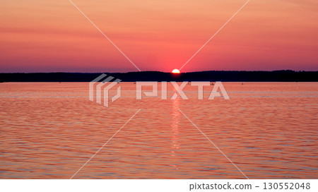 The sun sets behind a dark treeline, painting the sky pink. The sky's colors reflect on the calm lake, where people are boating and paddleboarding at dusk The sun sets behind a dark treeline, painting the sky pink. The sky's colors reflect on the calm lake, where people are boating and paddleboarding at dusk 130552048