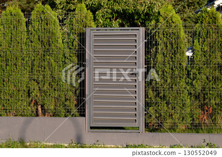 A modern gate of gray metal, with horizontal slats, stands closed in a fenced yard. The gate is flanked by green bushes and a metal mesh fence A modern gate of gray metal, with horizontal slats, stands closed in a fenced yard. The gate is flanked by green bushes and a metal mesh fence 130552049