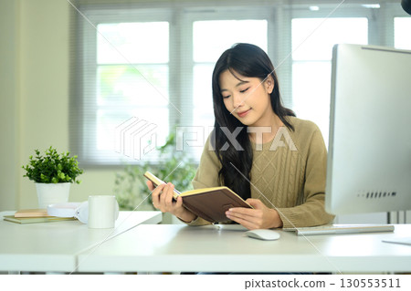 Young woman reading a notebook while sitting at desk with computer and coffee cup Young woman reading a notebook while sitting at desk with computer and coffee cup 130553511
