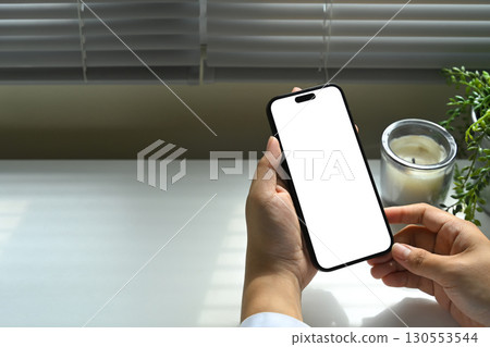 Hand holding smartphone with blank white screen, minimal desk setup with candle and plant. 130553544