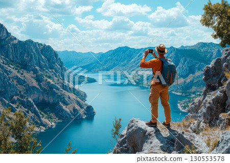 Backpacker stands on cliff, capturing stunning blue lake and rugged mountains with smartphone. Scenic view, nature exploration, travel photography. 130553578