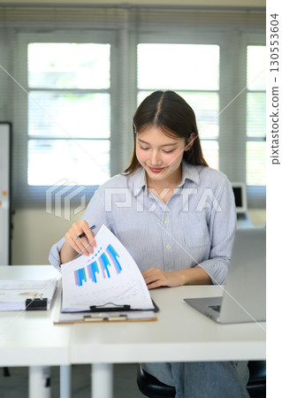 Businesswoman writing notes on paper while analyzing financial reports 130553604