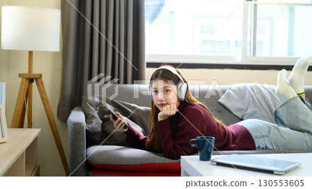 Young woman in headphones lying on sofa with smartphone, enjoying music at home Young woman in headphones lying on sofa with smartphone, enjoying music at home 130553605