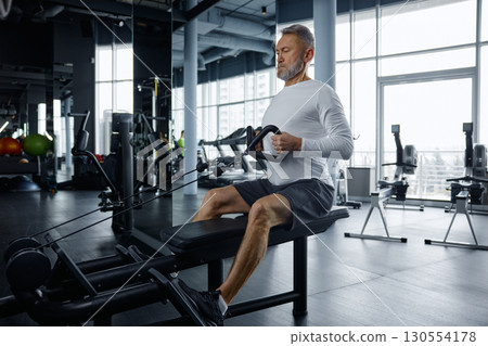 Senior man performs rowing exercise at modern gym Senior man performs rowing exercise at modern gym 130554178