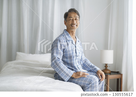 A man in his 60s sitting on a bed looking at the camera and smiling 130555617