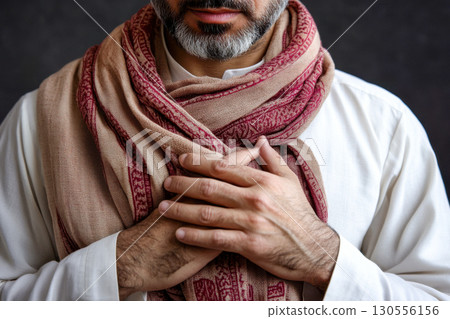 Close up portrait of mature man with grey beard wearing patterned beige red scarf. Hands clasped over chest show respect, comfort, gratitude, sincerity. 130556156