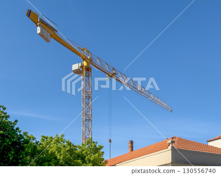 Tall construction crane above a red roofed building and green trees under a clear blue sky. urban 130556740