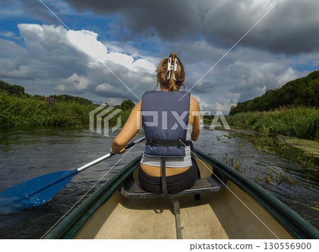 A young woman seen from behind canoeing in a river. Dark summer clouds in the background A young woman seen from behind canoeing in a river. Dark summer clouds in the background 130556900