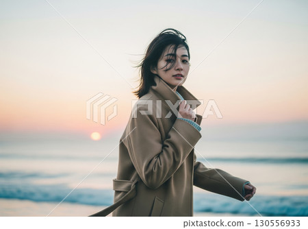 A woman wearing a beige coat standing on the beach at sunset in autumn A woman wearing a beige coat standing on the beach at sunset in autumn 130556933