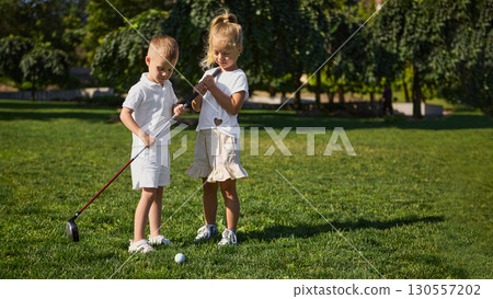 Boy and girl stand on grass preparing golf shot together 130557202