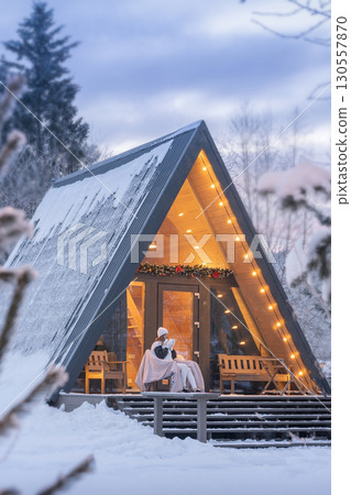 A magical winter morning outdoors in an A-frame cabin. A woman drinks tea or coffee on the porch of a house in a winter forest, late in the evening or early in the morning. 130557870