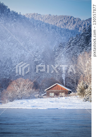 A cozy cabin situated in a picturesque snowy landscape, beautifully reflecting tranquility and winter beauty. Experience a Serene Winter Retreat by the Lake, Enveloped by Beautiful SnowCovered 130557877