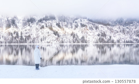 A serene winter landscape featuring snowcovered majestic mountains and their reflection, A small woman in a white jacket looks to the right at the majestic snow-capped mountains and their reflection A serene winter landscape featuring snowcovered majestic mountains and their reflection, A small woman in a white jacket looks to the right at the majestic snow-capped mountains and their reflection 130557878