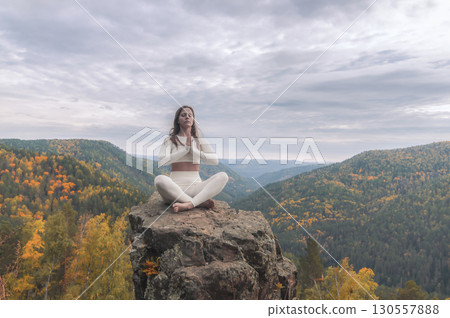 A white woman sits in the lotus position on a stone cliff, deep in meditation. Surrounded by breathtaking autumn landscapes, her serene presence harmonizes with the vibrant hues of the mountains. 130557888
