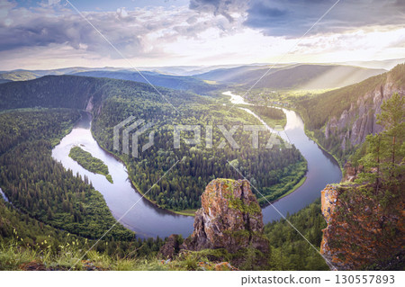 Stunning panorama showcases winding river flowing through vast green valleys surrounded by dense forest. Sunlight breaks through clouds, casting a warm glow over the landscape. Mana river Urman loop 130557893