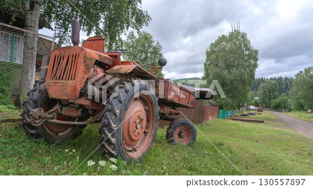 Old red rusty russian tractor t 16 parked beside gravel road in rural area surrounded by trees and old buildings during a cloudy afternoon 130557897
