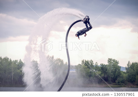 Flyboarding session showcases thrilling acrobatics over the water at a scenic lake on a cloudy afternoon. A man on a flyboard does acrobatic stunts over water Flyboarding session showcases thrilling acrobatics over the water at a scenic lake on a cloudy afternoon. A man on a flyboard does acrobatic stunts over water 130557901