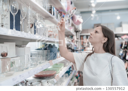 A woman stands in a store, thoughtfully assessing the quality of a wine glass she holds up to the light. Surrounded by shelves filled with various glassware, she appears focused on making A woman stands in a store, thoughtfully assessing the quality of a wine glass she holds up to the light. Surrounded by shelves filled with various glassware, she appears focused on making 130557912