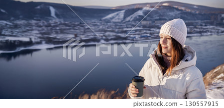 A Woman Delightfully Enjoying Her Cup of Coffee by a Scenic Winter River, Fully Embracing the Moment A winter woman in a white jacket enjoys the beautiful view of the mountains and the blue river with 130557915