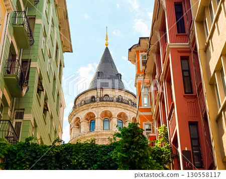 Close view of Galata Tower framed by colorful buildings and greenery in Istanbul. Historical heritage, architectural design, and cultural landmark symbolizing urban identity. 130558117