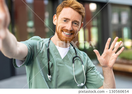 Satisfied man doctor in scrubs waving hand while taking pov selfie 130560654