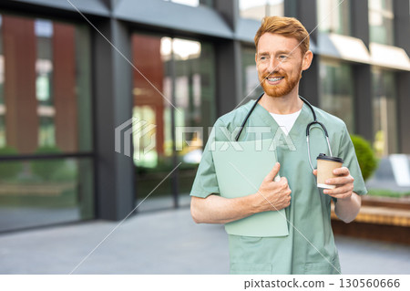 Man physician in scrubs standing near hospital with cup and folder Man physician in scrubs standing near hospital with cup and folder 130560666