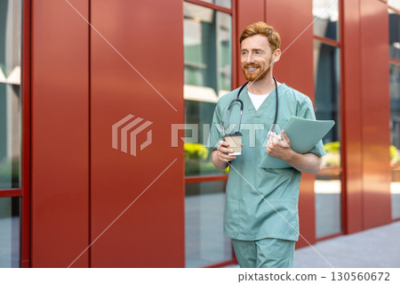 Male medic carrying coffee cup and documents near hospital building 130560672