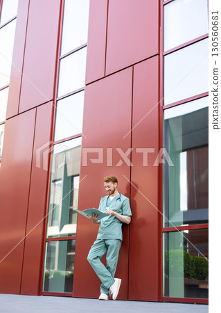 Bearded man in scrubs standing against wall reading documents 130560681