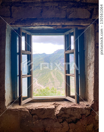 Old wooden window in stone building opens to reveal lush green mountains under cloudy sky 130561064
