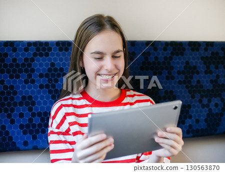 Teen girl wearing striped shirt smiles as she watches something on her tablet while seated in train 130563870