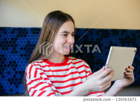 Teen girl wearing striped shirt smiles as she watches something on her tablet while seated in train 130563871