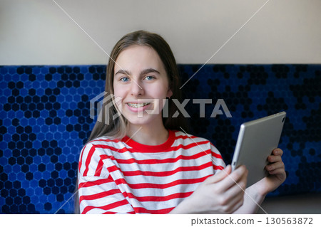 Teen girl wearing striped shirt smiles as she watches something on her tablet while seated in train 130563872