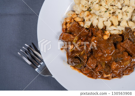 Delicious hungarian goulash with spaetzle on white plate resting on gray tablecloth with fork Delicious hungarian goulash with spaetzle on white plate resting on gray tablecloth with fork 130563908