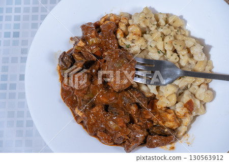 Fork taking a bite of beef goulash with spaetzle on white plate 130563912