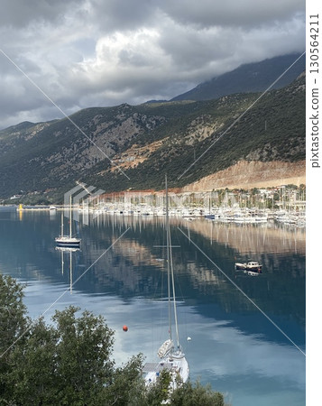 Marina with sailboats and reflections on calm water, surrounded by mountains under cloudy sky 130564211