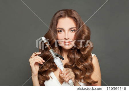 Attractive woman with long wavy hairstyle using hair curling tongs iron, indoor portrait. 130564546