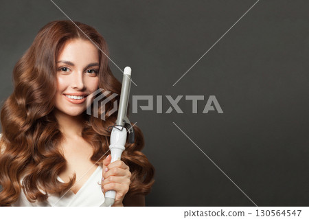 Gorgeous woman with long wavy hairstyle using hair curling tongs iron, indoor portrait. Gorgeous woman with long wavy hairstyle using hair curling tongs iron, indoor portrait. 130564547