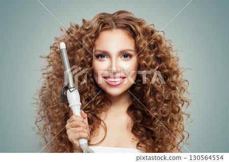 Smiling woman with long wavy hairstyle using hair curling tongs iron, indoor portrait.  130564554