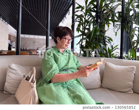 Middle-aged woman in green dress reading book on sofa. Knowledge, leisure, and personal time in modern lifestyle. Middle-aged woman in green dress reading book on sofa. Knowledge, leisure, and personal time in modern lifestyle. 130565852