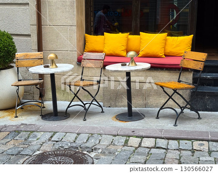 Outdoor cafe with wooden chairs, marble tables, and bright yellow pillows. Hospitality, comfort, and lifestyle in an urban social setting. 130566027