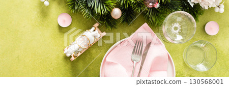 Top view of an elegant Christmas table with an empty plate, folded pink napkin and cutlery on a vibrant green background. Spruse branches and pink tree decorations add seasonal charm, banner Top view of an elegant Christmas table with an empty plate, folded pink napkin and cutlery on a vibrant green background. Spruse branches and pink tree decorations add seasonal charm, banner 130568001