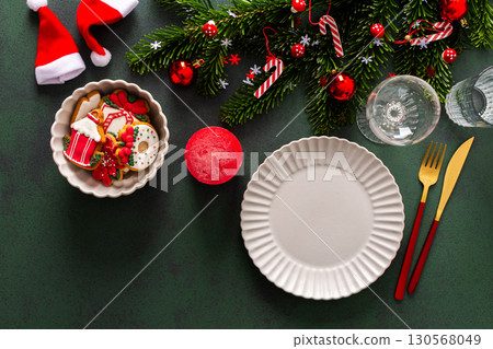A Christmas table setting featuring an empty plate, napkin and gingerbread ,cutlery and a candle on a deep green background. Spruse branches and red tree decorations create a festive atmosphere A Christmas table setting featuring an empty plate, napkin and gingerbread ,cutlery and a candle on a deep green background. Spruse branches and red tree decorations create a festive atmosphere 130568049