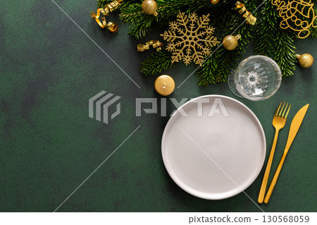 A Christmas table setting with an empty plate and cutlery on a green background. Spruse branches and golden tree decorations add elegance and festive cheer to the scene A Christmas table setting with an empty plate and cutlery on a green background. Spruse branches and golden tree decorations add elegance and festive cheer to the scene 130568059