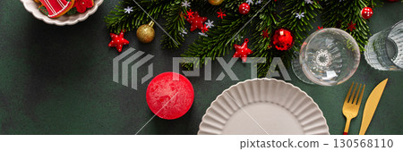 A cozy Christmas table with an empty plate, cutlery and candle on a rich green background. Red Christmas tree decorations and spruse branches complete the festive look, banner 130568110