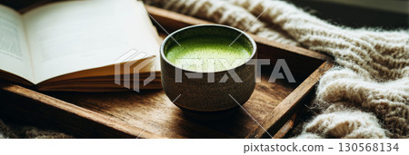 A cup of frothy matcha tea sits on a wooden tray beside a cozy knit sweater and an open book, creating a peaceful atmosphere perfect for relaxation and enjoyment, banner 130568134