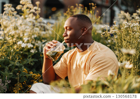 A man savors a matcha beverage while sitting among vibrant flowers and greenery. The warm sunlight creates a tranquil ambiance, enhancing the peaceful moment of relaxation A man savors a matcha beverage while sitting among vibrant flowers and greenery. The warm sunlight creates a tranquil ambiance, enhancing the peaceful moment of relaxation 130568178
